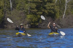 Two people kayaking on a river lined with green trees, in Iron County, Michigan, on a nice day.