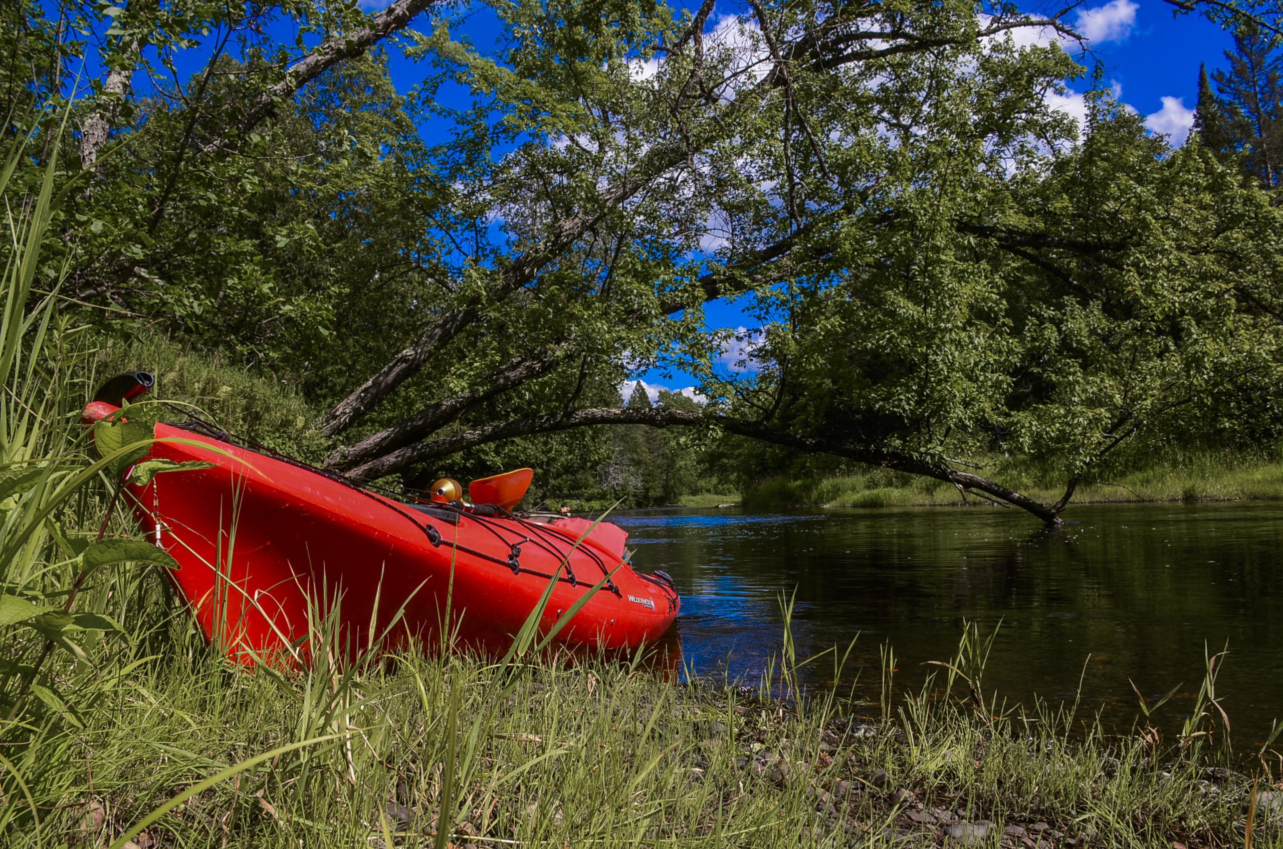 A red kayak resting on the banks of a river in Iron County, Michigan, on a sunny day.