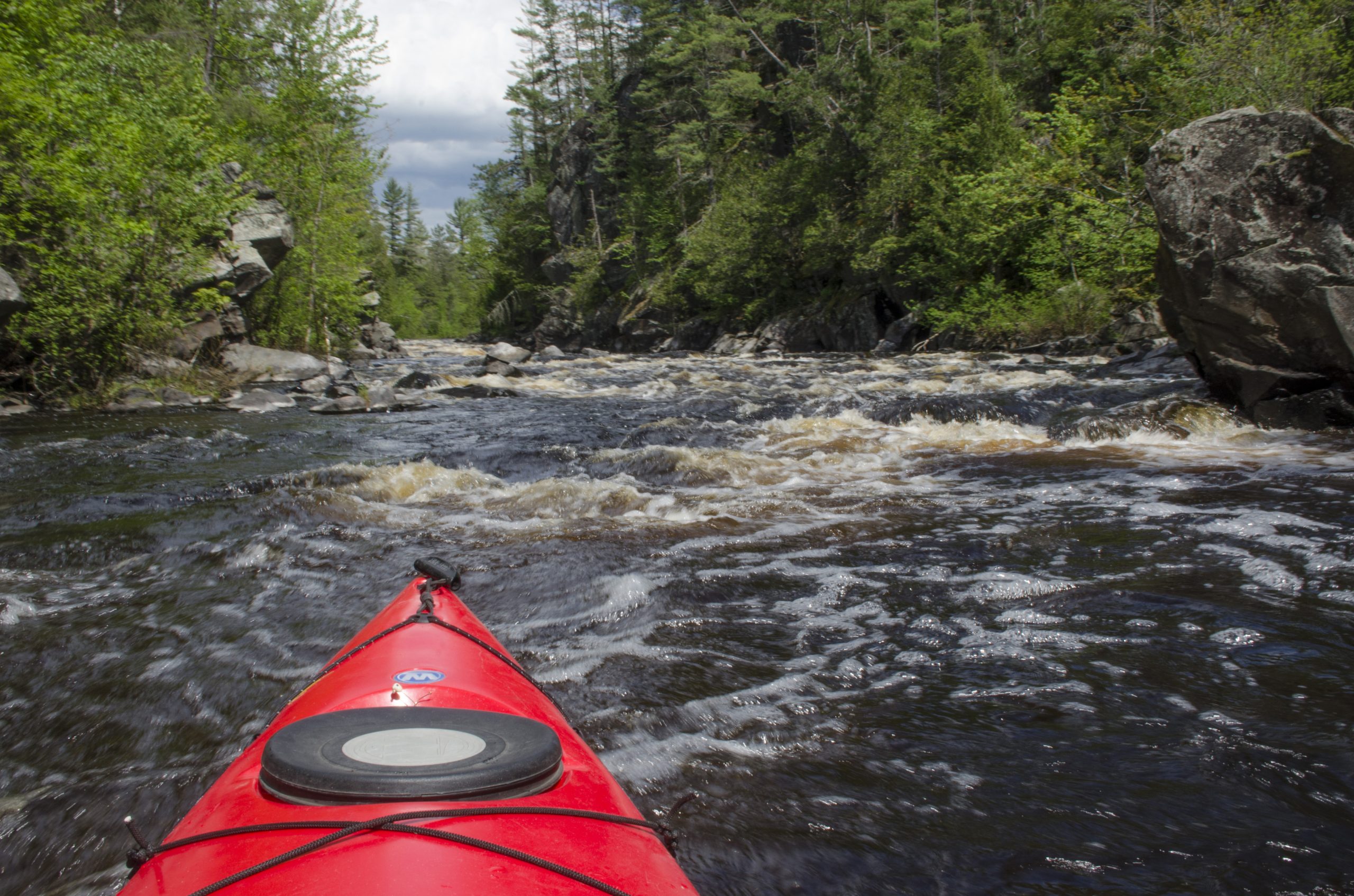 The red bow of a kayak heading into river rapids between green banks