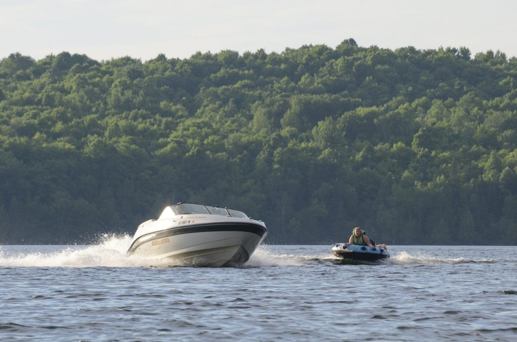 A boat pulling a person on a tube across a lake in Iron County, Michigan on a nice summer day. The lake is lined by trees.
