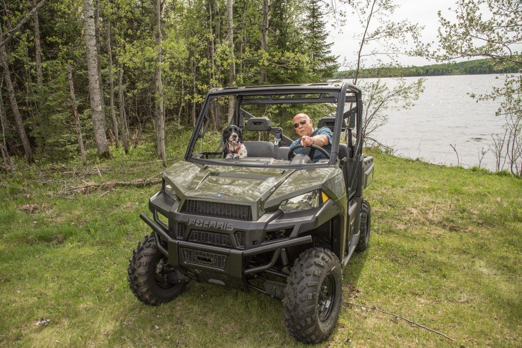 A man and his dog sitting in a parked side-by-side ATV in front of a lake in Iron County, Michigan, on a summer day, surrounded by lush green trees.