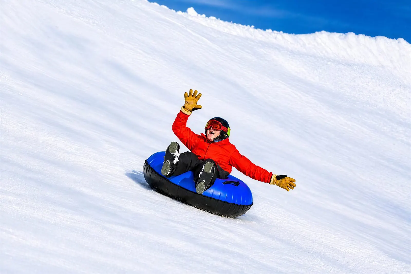 A child in a red jacket waving while snow tubing down a groomed hill on a blue tube