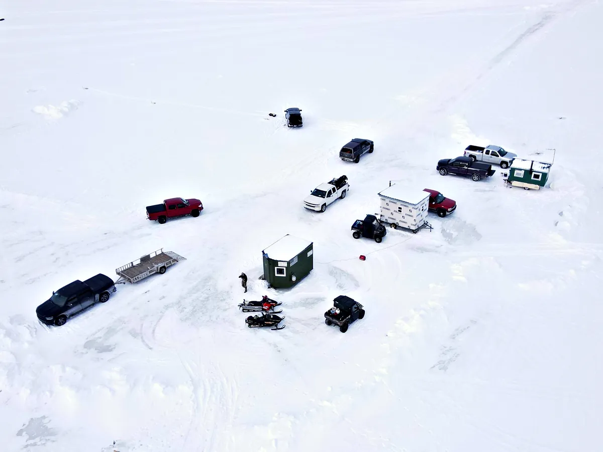 Aerial view of an Iron County ice fishing camp: trucks, snowmobiles, a side-by-side and wooden ice shanties on a frozen lake