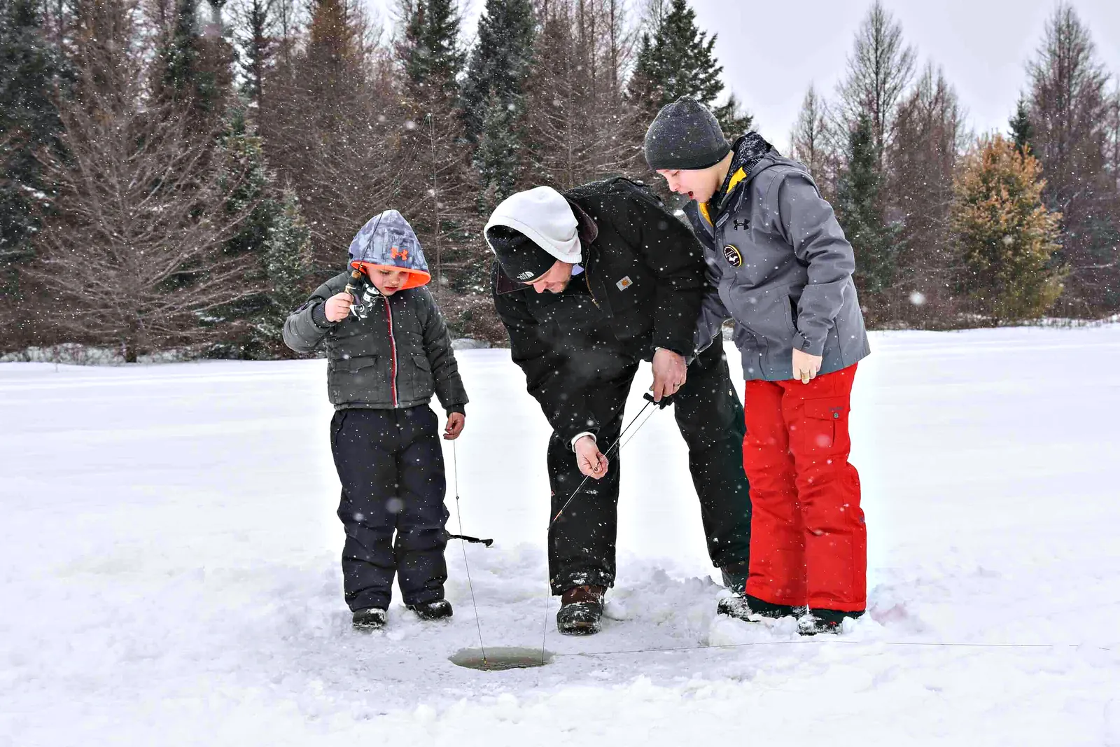 A dad showing two boys how to jig through a hole in the ice as snow falls