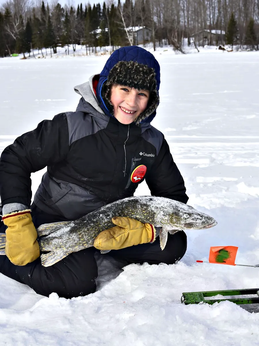 A smiling boy in a trapper hat holding a northern pike on the ice beside a tip-up with its orange flag